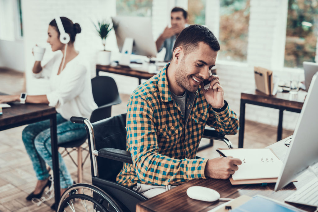 Male wheelchair user sits at office desk, writing notes while talking on the phone smiling. Background shows a women sitting at a desk with headphones on drinking from a cup and another male sitting at a computer.