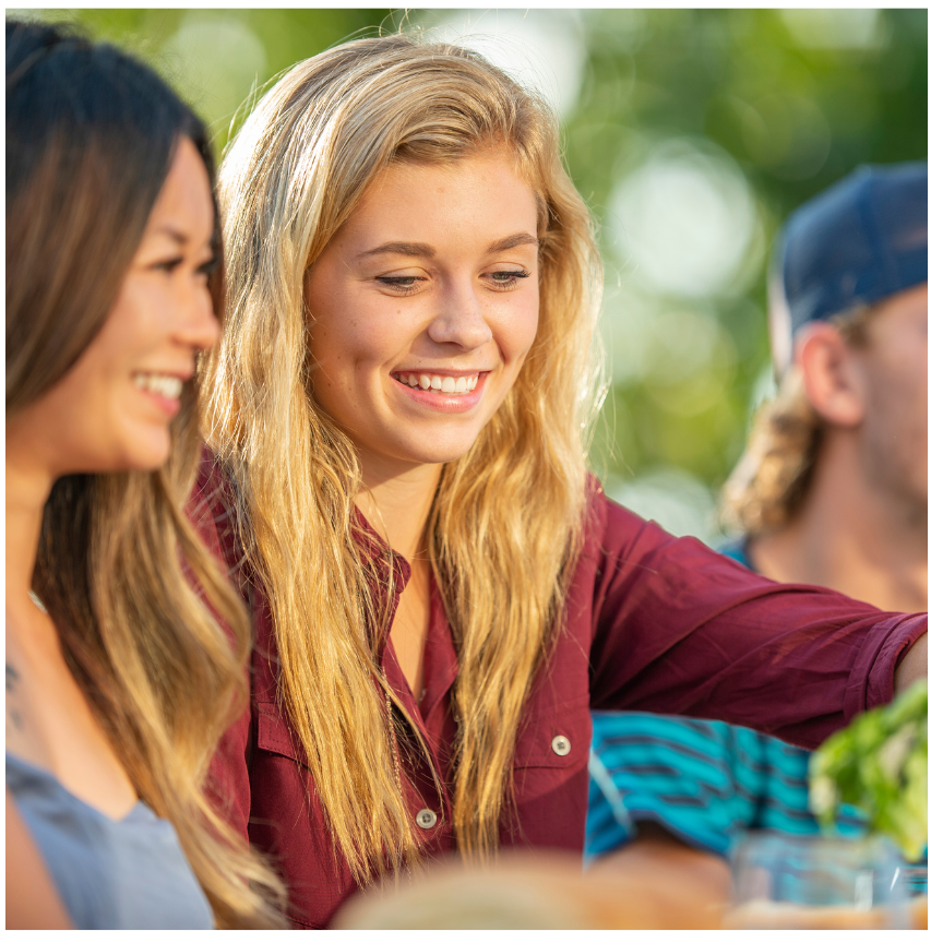 Tw young women in shot with one in focus. Woman is looking down and smiling. She has long blonde hair and is wearing a maroon shirt.