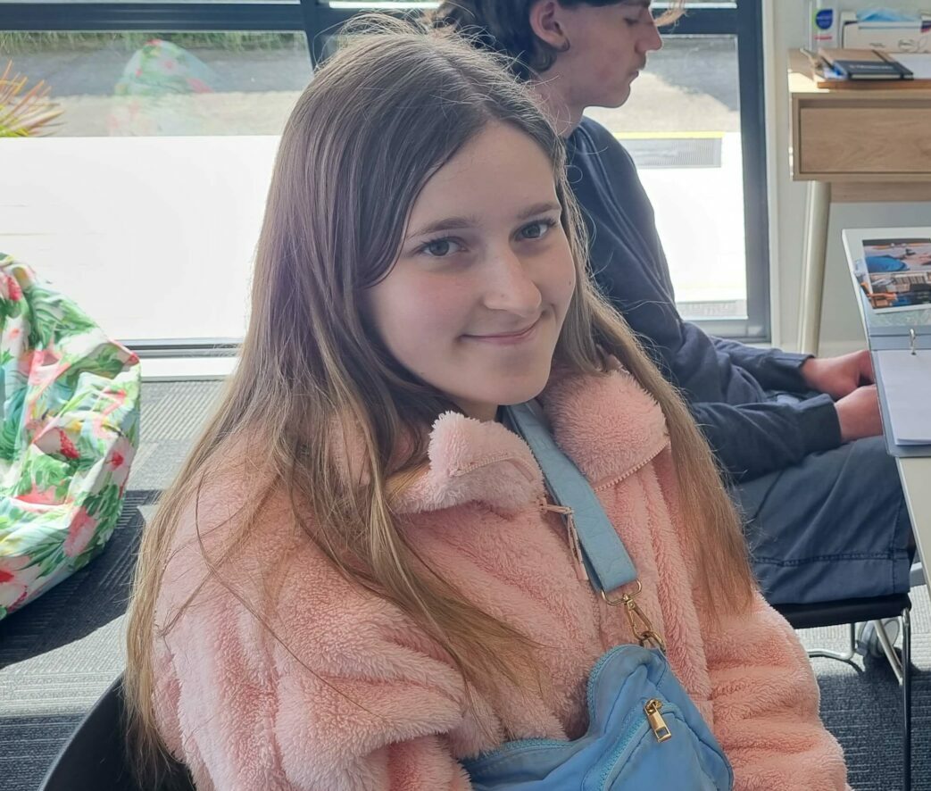 Momenta client Madeleine sitting down wearing a pink zip up jumper and blue bag over the shoulder.