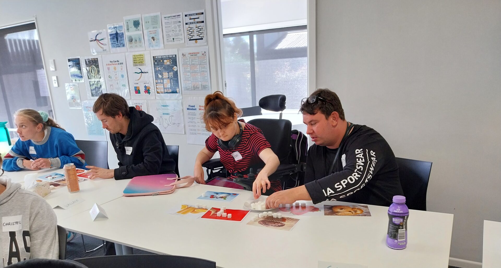 Four Momenta clients sitting at a table working through course materials. One wearing a blue jumper, one wearing a black hoodie and one in a black wheelchair wearing a striped tshirt and one wearin ga black tshirt with writing on it.