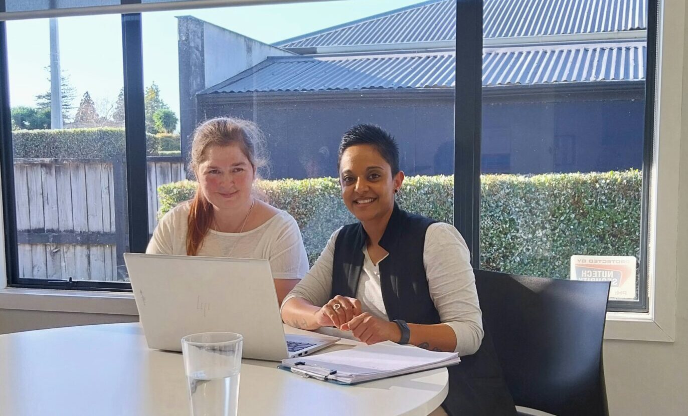 Amy sitting on the left at a table wearing a white tshirt. Beena sitting on the right wearing a white tshirt and black vest.